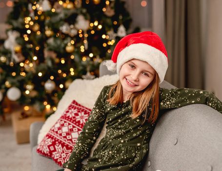 Little girl in santa hat hugging teddy bear while sitting on sofa near christmas tree at home