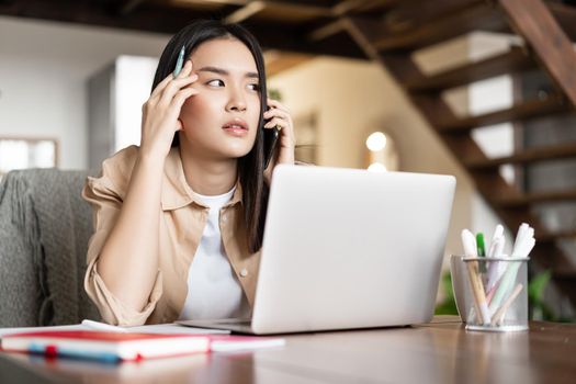 Asian girl answer phone call, looking thoughtful while communicating on phone. Young woman working from home on laptop.