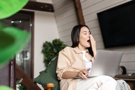 Tired asian girl yawns during work on laptop, sits at home, works remotely from her living room.