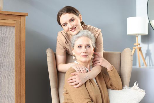 Happy daughter hugging older mother, standing behind chair in living room, enjoying tender moment at home