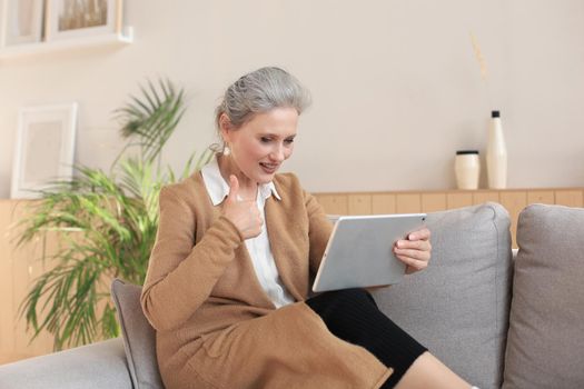 Cheerful middle aged woman sitting on sofa, using computer tablet apps, looking at screen,reading good news in social network, shopping or chatting online