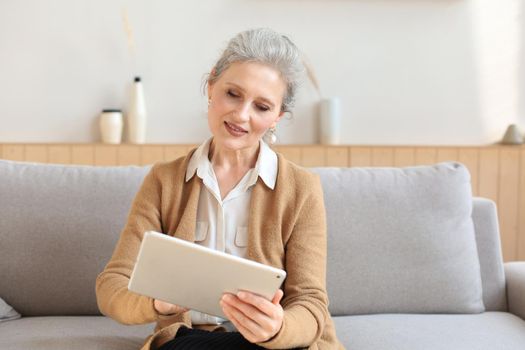 Cheerful middle aged woman sitting on sofa, using computer tablet apps, looking at screen,reading good news in social network, shopping or chatting online