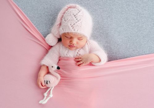 Cute newborn baby girl in knitted hat sleeping on grey stylized background and holding flamingo toy in her hands. Adorable infant child napping under pink blanket during studio photoshoot