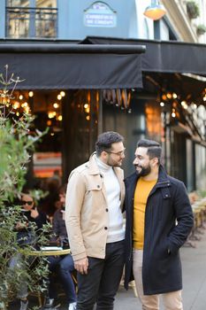Two young gays standing near street cafe and smiling. Concept of same sex couple and male friendship.