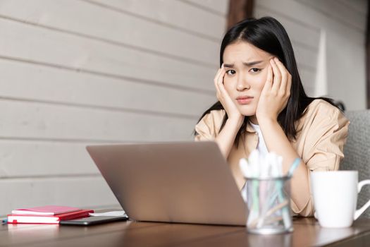 Gloomy asian female student sitting bored with laptop at home, tired or bored of studying, having online classes remote indoors, doing homework or working.