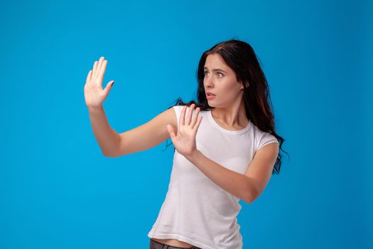 Studio portrait of a young confused woman in white t-shirt against a blue wall background. She act like she is asking someone to stop. People sincere emotions, lifestyle concept. Mock up copy space.