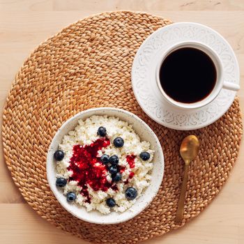 breakfast with white bowl of homemade curd with jam, raspberries, blueberries and cup of coffee on brown wooden table, top view, close up