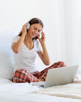 Young smiling woman sitting on bed in bedroom and listening to music or watching movie on laptop. Distance learning, surfing Internet in morning after waking up