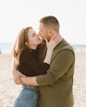 Young adult couple standing on beach and looking each other. Handsome smiling man embracing beautiful woman full of love. Millennial male and female posing on coast on sunny day and looking away