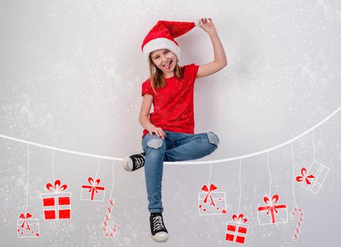 Little girl in santa hat sitting on chalk drawn thread with hanging christmas presents on white wall background