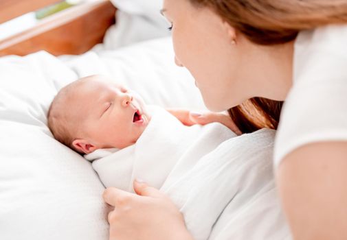 Young mother looking at her newborn daughter swaddled in white sheets at home sleeping with mouth open. Portrait of girl mom with her infant child napping in the bed