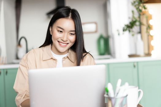Smiling asian girl using laptop, working from home on computer pc, sitting in kitchen and studying, doing homework.