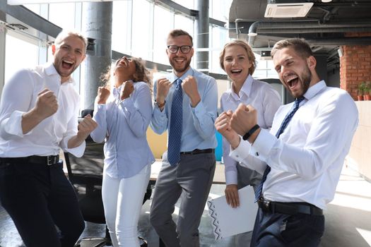 Five happy modern business people are keeping arms raised and expressing joyful while standing in large office.