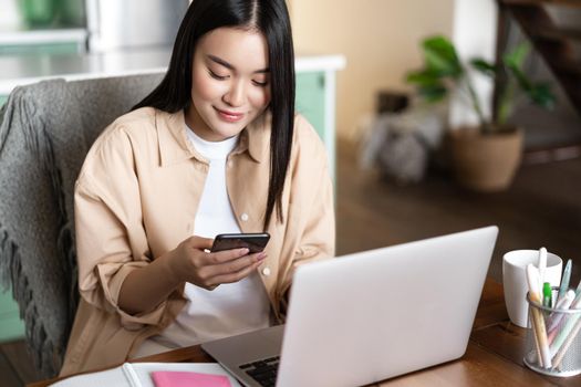 Image of asian woman checking phone messages, working on laptop computer from home, waiting for work conference, video chat.