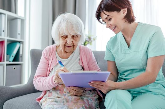 Elderly woman and nurse sign documents at home. Healthcare worker girl cares about senior female person indoors