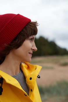 One with nature. Crop closeup portrait of mature beautiful woman in bright yellow jacket and red hat on shore of North sea on autumn day. Mood Scandinavian weather.