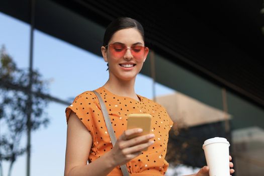 Portrait fashion woman in yellow dress in sunglasses walking on street and holding smartphone in her hand