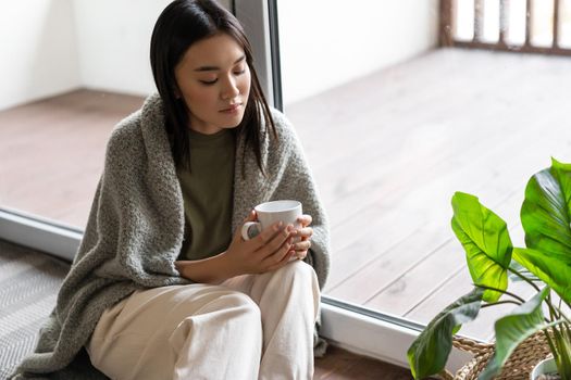 Young sad asian woman sitting on floor near balcony and looking at tea mug thoughtful.