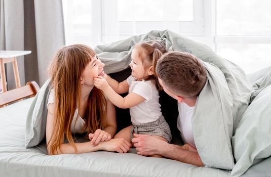 Beautiful family under the blanket in the bed. Young parents playing with small daughter under coverlet and smiling in sunny room. Child touching her mother face and father hugging the kid