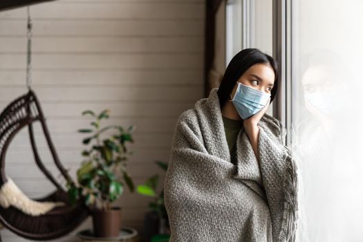 Sad sick asian girl on quarantine, standing near window at home and looking outside, wearing medical face mask from covid.