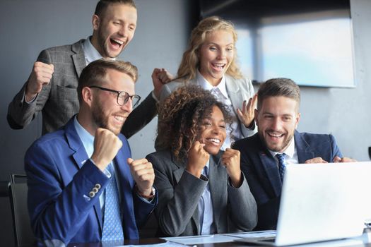 Five happy modern business people are keeping arms raised and expressing joyful while sitting near laptop in the office