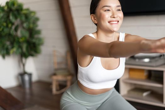 Young asian fitness woman with healthy fit body, doing squats, morning workout, wearing activewear, standing at home in living room and having training session.