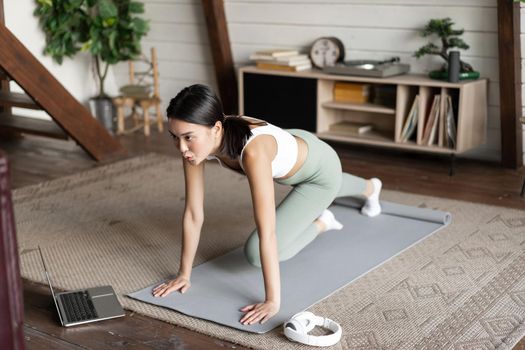 Image of young asian girl doing workout at home, following fitness instructor online on laptop, doing exercises in living room, wearing activewear.