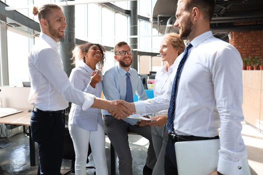 Two smiling businessmen are shaking hands while being in office together with their colleagues.