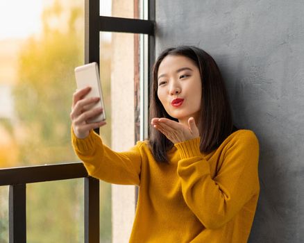 Young Asian woman takeing selfie and blowing kiss. Beautiful teenage girl with bright red lips siting on window and communicating via video call with boyfriend