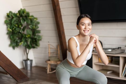 Young asian fitness woman with healthy fit body, doing squats, morning workout, wearing activewear, standing at home in living room and having training session.