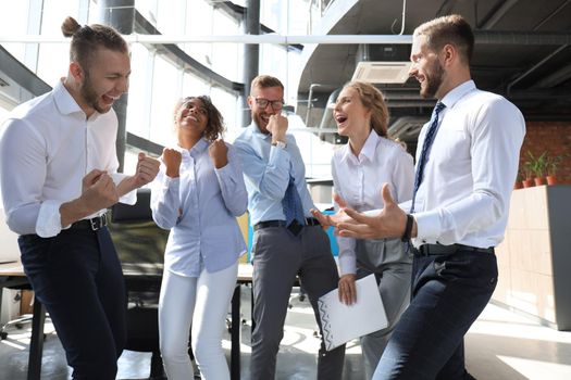 Five happy modern business people are keeping arms raised and expressing joyful while standing in large office.