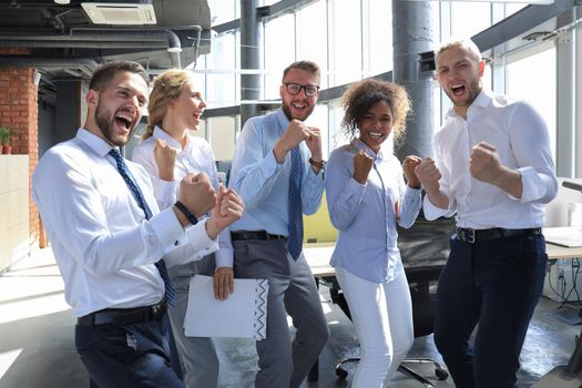 Five happy modern business people are keeping arms raised and expressing joyful while standing in large office.