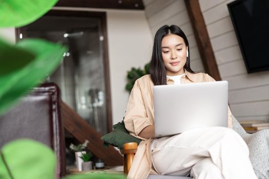 Young asian girl watching webinar on laptop. Girl relaxing with computer in hands at home, sitting on chair resting.