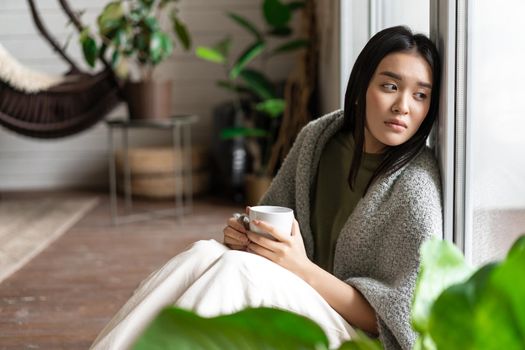 Sad asian woman looking outside window, sitting on floor with miserable upset face, drinking coffee at home.