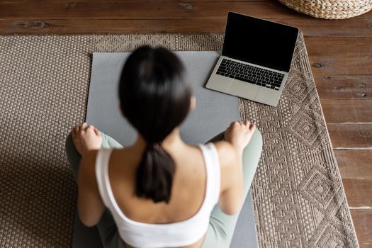 Mindfulness and meditation concept. Rear view of young asian woman meditating at home, following online course on laptop, sitting in living room on floor mat.