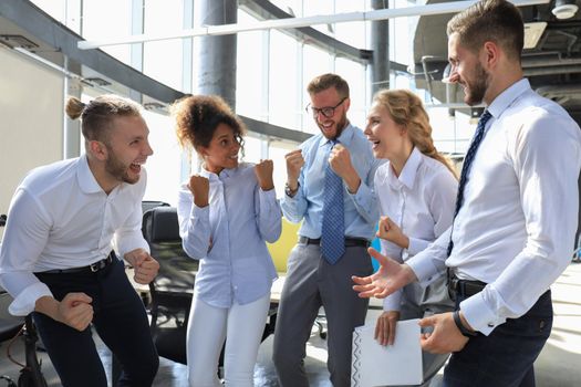 Five happy modern business people are keeping arms raised and expressing joyful while standing in large office.