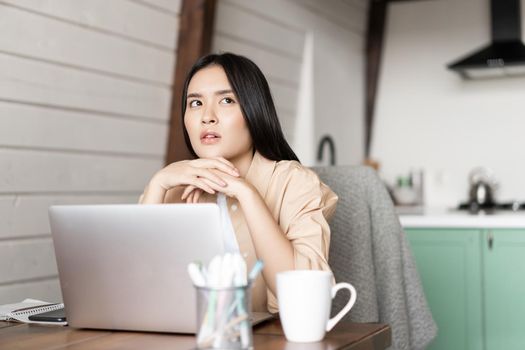 Portrait of thoughtful asian girl working on laptop from home. thinking while studying online, listening webinar on computer.