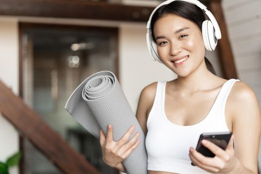 Smiling asian girl in headphones, listening music at home and workout with floor mat, holding mobile phone, fitness exercises in living room.