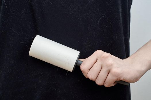 An unrecognizable woman cleans a black t-shirt with a clothes roller. A hand holding a cleaning tool against a gray wall