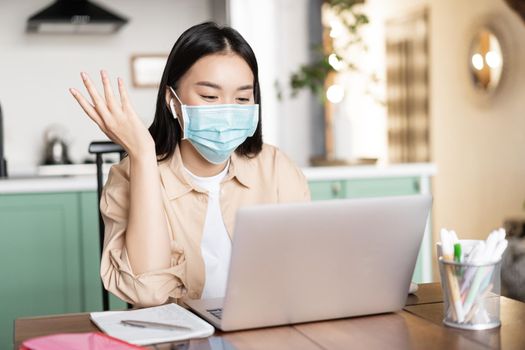 Cheerful asian woman working from home, communicating on laptop, wearing medical mask. Girl on quarantine studying via online classes.