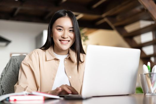 Smiling asian girl doing homework, studying online on laptop from home. Young woman working on computer pc indoors, remote work and e-learning concept.