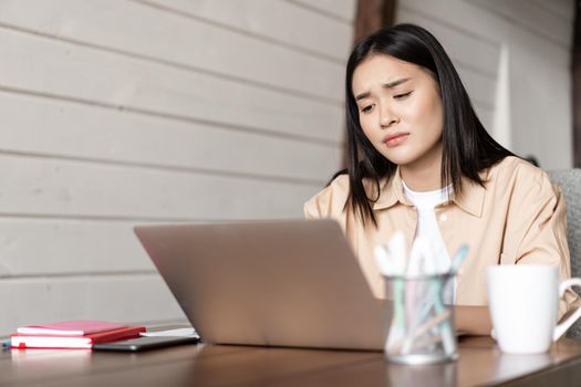 Sad and tired asian girl looking disappointed at laptop screen, bored of studying, watching something boring on computer.