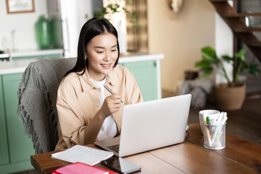 Smiling asian girl studying at home on laptop, listening webinar and taking notes, writing down homework with computer.