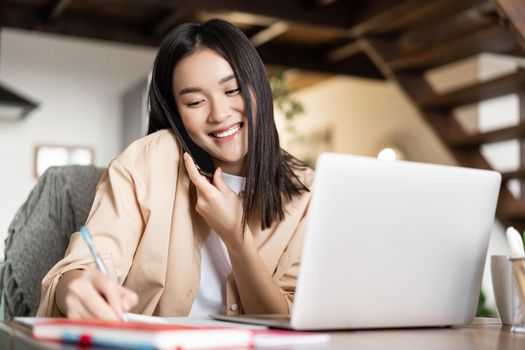 Smiling asian woman working from home, taking notes in notebook, talking on cellphone and writing down information, using laptop.