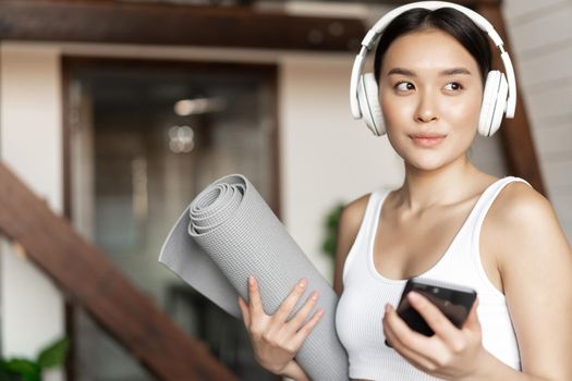 Young asian woman standing at home in headphones, listening music and going workout on floor mat, practice yoga or meditate in living room.