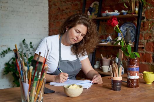 Artisan woman writing n notebook, planning day in diary. Beautiful business female entrepreneur with long curly hair sits at workshop in chair and thinking about working on projects