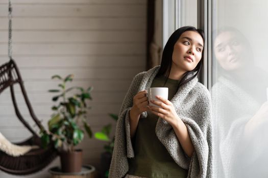 Romantic asian woman wrapped in blanket, leaning on window and looking outside, drinking hot coffee. Lifestyle concept