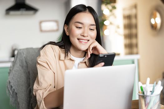 Smiling asian woman working remotely from home, using laptop and mobile phone, sitting at table with computer.