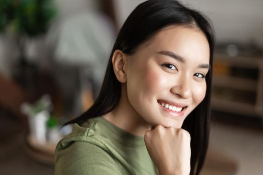 Portrait of young asian woman with clean glowing skin, looking and smiling at camera, sitting at home alone in living room. Lifestyle concept.