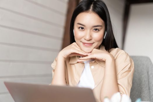 Portrait of asian woman in earphones working on laptop and smiling, looking happy at camera, studying or learning online course on computer.